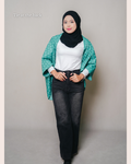 Woman wearing a green ponco like cardigan with Sabah pattern on it, tie at the back, white shirt, and black jeans against a plain background